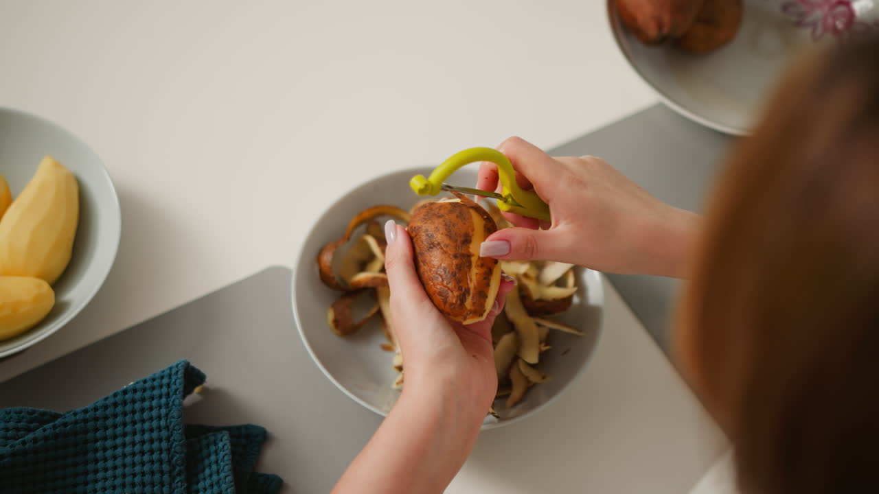 Top down view of cook peeling brown potato with yellow peeler over plate of discarded skins, surrounded by peeled potatoes, unpeeled potatoes, and kitchen towel on clean kitchen countertop