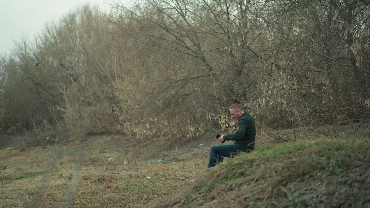 A close view of a man in a green jacket sitting alone in a secluded, wooded area, seemingly deep in thought or contemplation, holding a cell phone and piston
