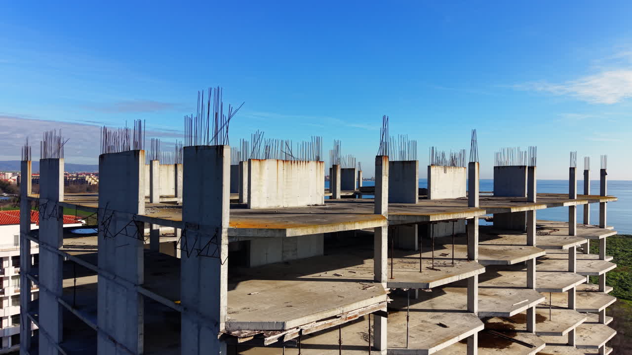 High angle shot showing the top floors of an abandoned or unfinished building with many concrete pillars and exposed rebar near the sea. Aerial view