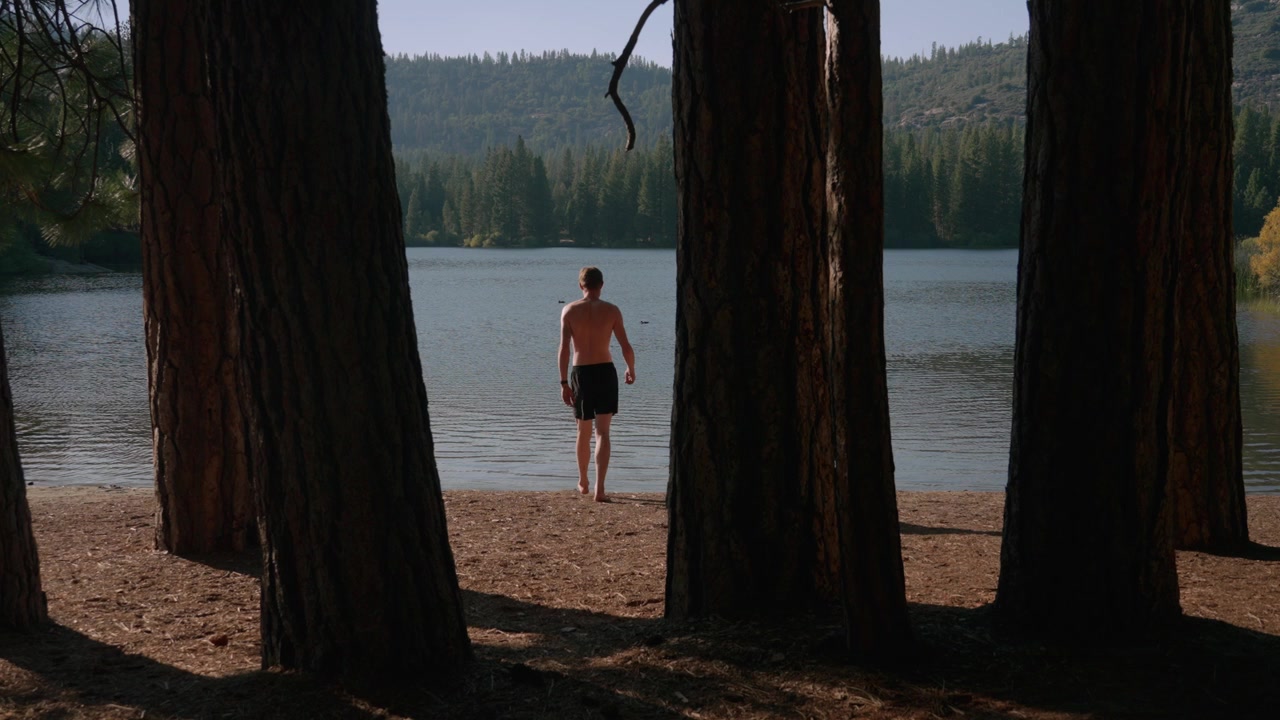 A shirtless man walks toward a calm lake between tall pine trees to go for a swim.