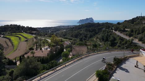 drone footage showing forested countryside and winding scenic hillside roads in calpe, spain, with a cyclist riding around a corner in the foreground, and the horizon and mediterranean sea behind