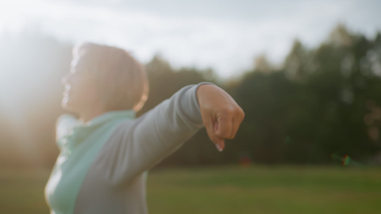 Angle view of lady wearing ash and mint tracksuit performing hand rotation exercise during morning walk in open green field with sunlight streaming across natural environment surrounded by trees