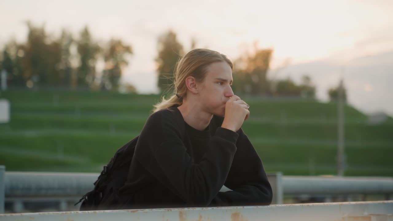 Jugendliche Person ruht sich aus. Ein Teenager pausiert friedlich an einem Zaun. Ein junger Mann entspannt sich ruhig am Parkzaun in der Abenddämmerung. Eine junge Person macht eine friedliche Pause, während sie sich in den Sonnenuntergangsstunden an ein Geländer lehnt.