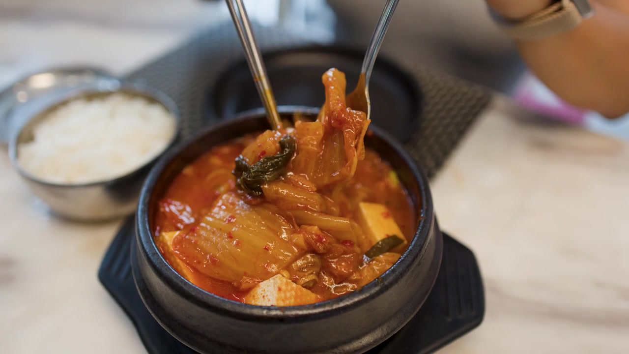Gambar close-up seseorang yang menggunakan sumpit logam dan sendok untuk menyantap sup kimchi Korea pedas