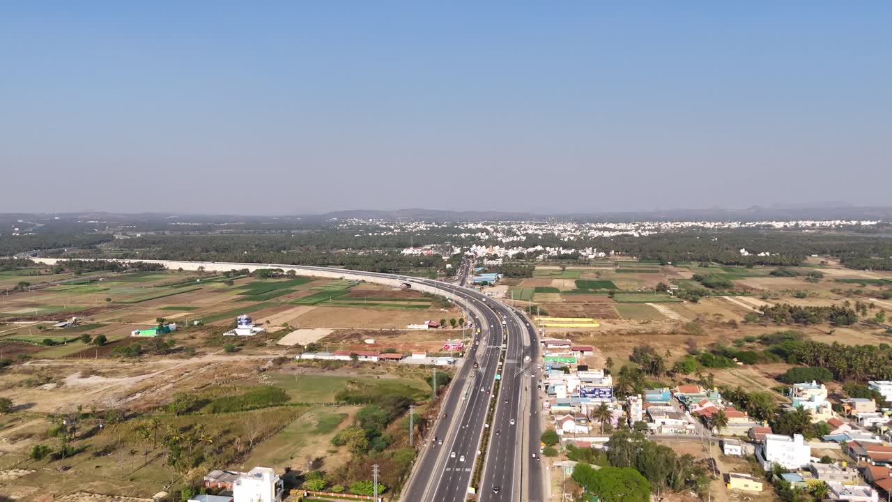 Aerial view: Multi-lane Chennai - Bengaluru highway, busy with traffic, runs alongside palm groves and a large lake. Residential areas and development blend with natural elements under a clear sky