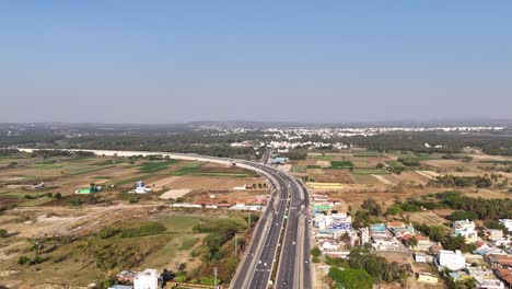 aerial view: multi-lane chennai - bengaluru highway, busy with traffic, runs alongside palm groves and a large lake. residential areas and development blend with natural elements under a clear sky