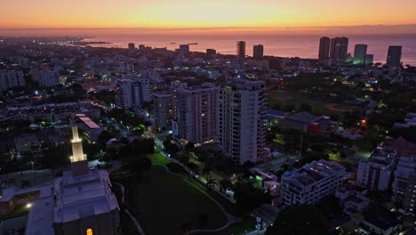 aerial shot of santo domingo at sunrise, moving backward to reveal the skyline, caribbean sea, and the warm golden sunlight illuminating the buildings and horizon in a peaceful morning atmosphere