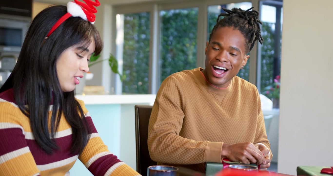 Diverse couple at kitchen table for Christmas, man untying ribbon while woman holding mug
