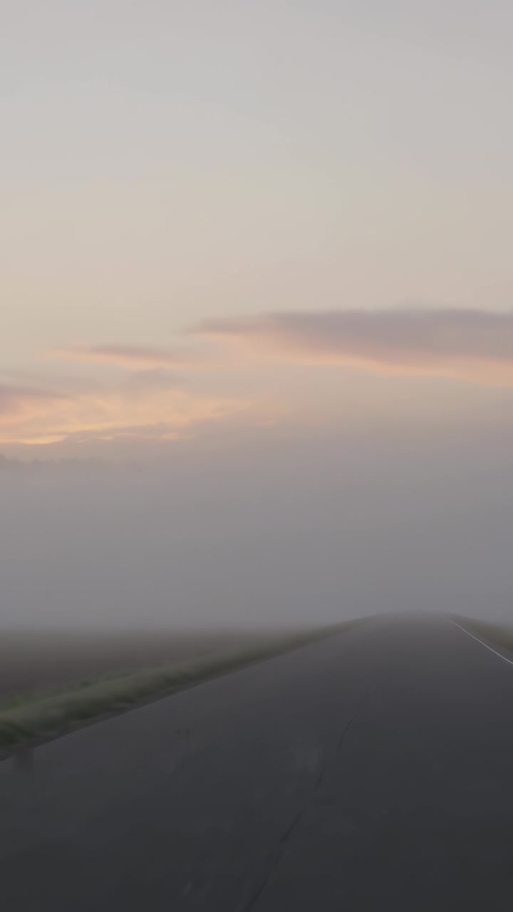 Vertical video: Moving camera revealing two-lane road entering low mist over plains, pink sky