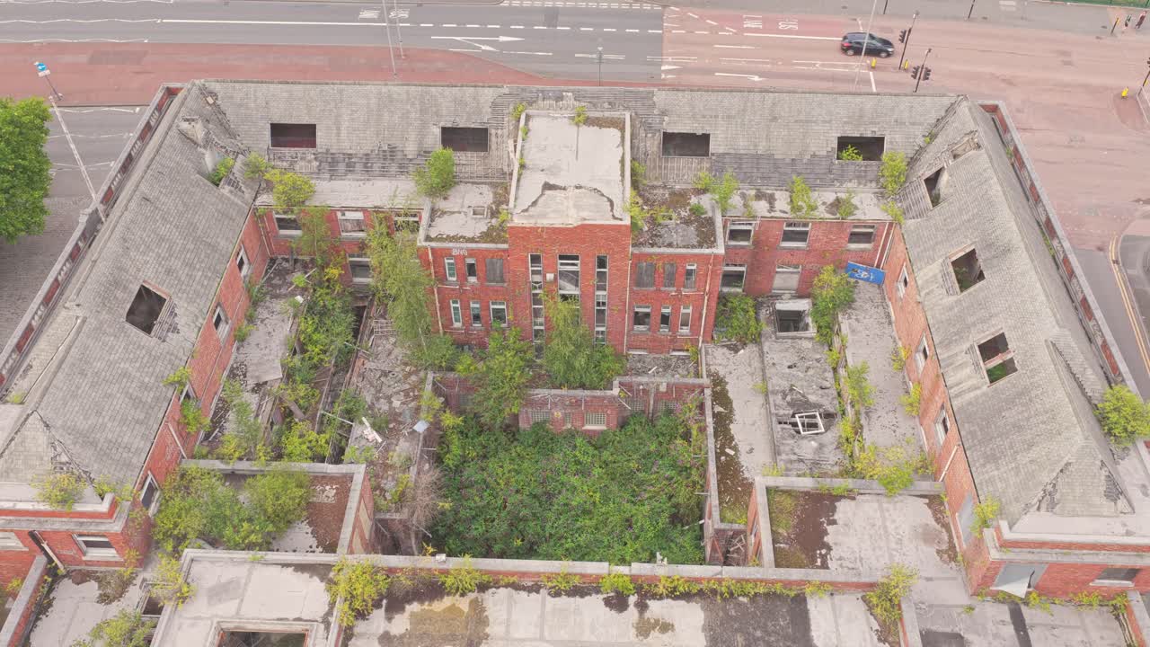 Drone ascending shot of the abandoned Salford Crescent Police Station on The Crescent (A6), with vegetation overtaking its courtyard and broken structures, symbolizing urban decay