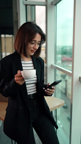 good-looking caucasian brunette drinking coffee and using gadget. lady on a coffee break in the office. vertical video.
