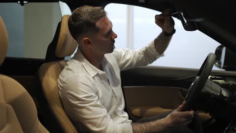 cliente con camisa blanca sentado en el salón de automóviles, toca el espejo, quiere comprar un automóvil nuevo en la sala de exposición