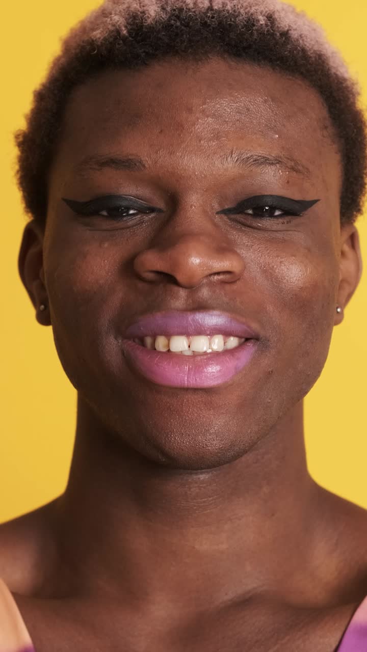 African trans person with colorful dress and make up smiling at the camera on yellow