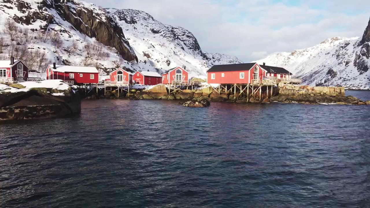 Luftfoto av Nusfjords tradisjonelle røde rorbuer og turkist arktisk vann i et snørikt vinterlandskap, Lofoten, Norge. Ikonisk landskap fra en skandinavisk fiskelandsby.