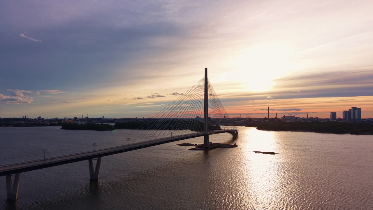 Ponte strallato Kruunuvuorensilta a Helsinki, Finlandia, che attraversa acque calme al tramonto con un cielo caldo e dorato, skyline della città ed edifici industriali visibili sullo sfondo.