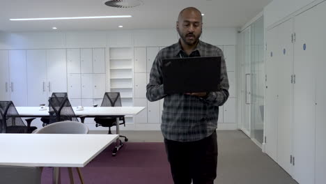 an asian indian man pacing around an empty office holding his laptop computer while talking to colleagues during an online conference call meeting