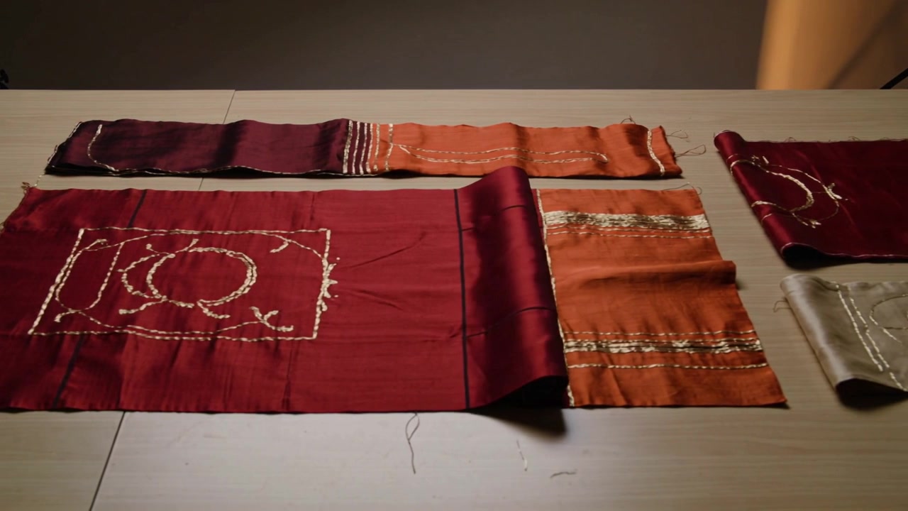 Unrolling large maroon cloth with stitched motif on wood table after roll release to reveal motif
