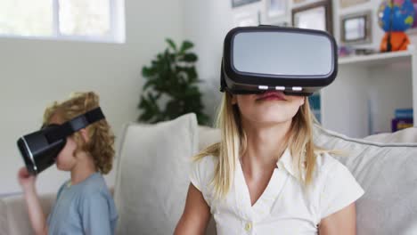 Caucasian-brother-and-sister-gesturing-while-using-vr-headset-sitting-on-the-couch-at-home