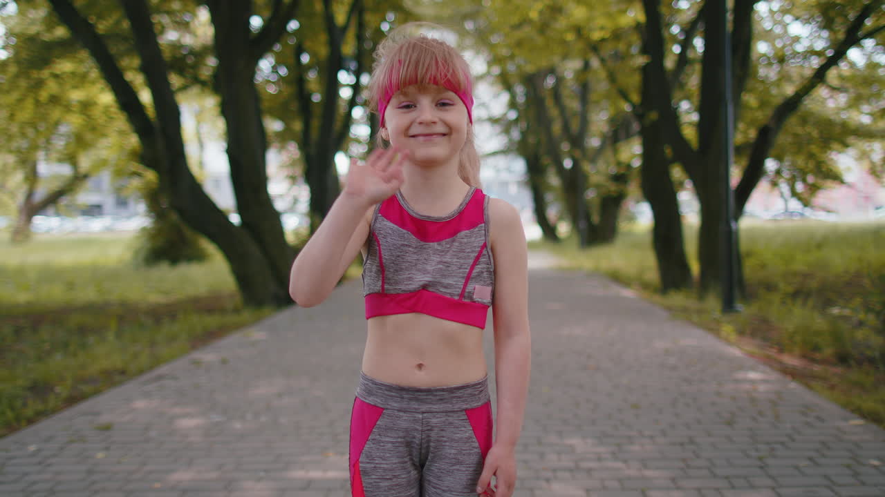 Atletisk og sprek småbarnsjente som løper, vinker hei med håndflaten og hilser med et gjestfritt, vennlig smil.