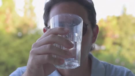 hombre con gafas de sol bebiendo agua y relajándose en el jardín con vegetación exuberante en el fondo