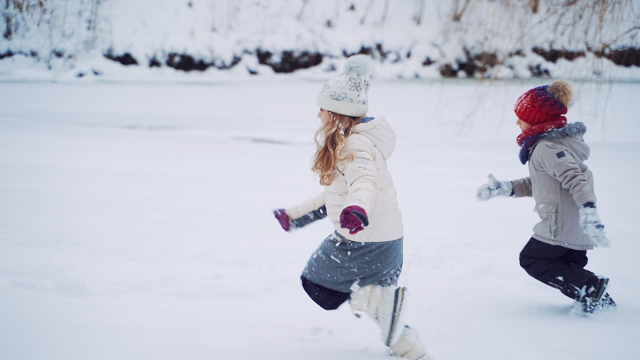 Side view of two children in warm clothes and caps are running on the snowy background. Active kids are playing in winter near the forest. Slow motion.