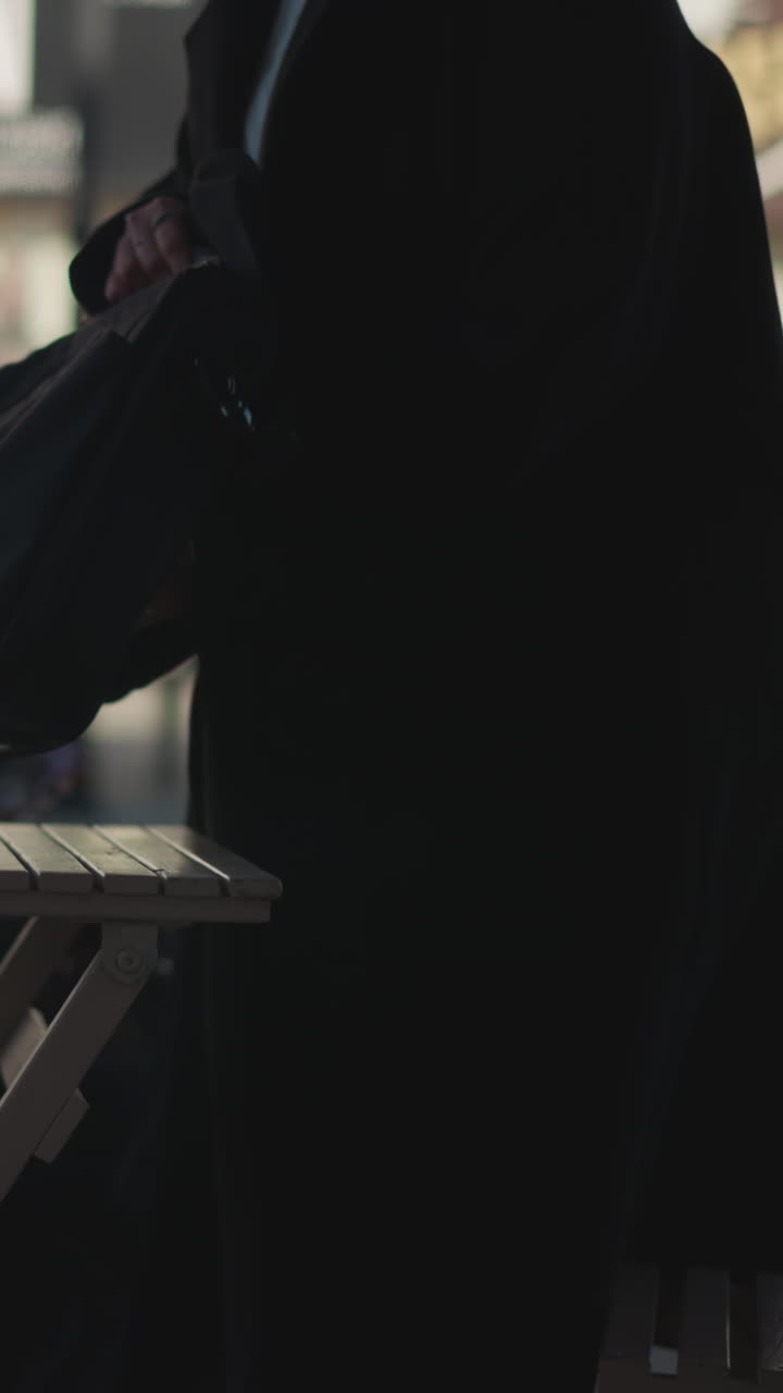 una trabajadora remota se prepara para sentarse en una cafetería al aire libre, colocando su bolsa en una mesa, el fondo borroso destaca elementos urbanos, incluidos edificios, postes y flores