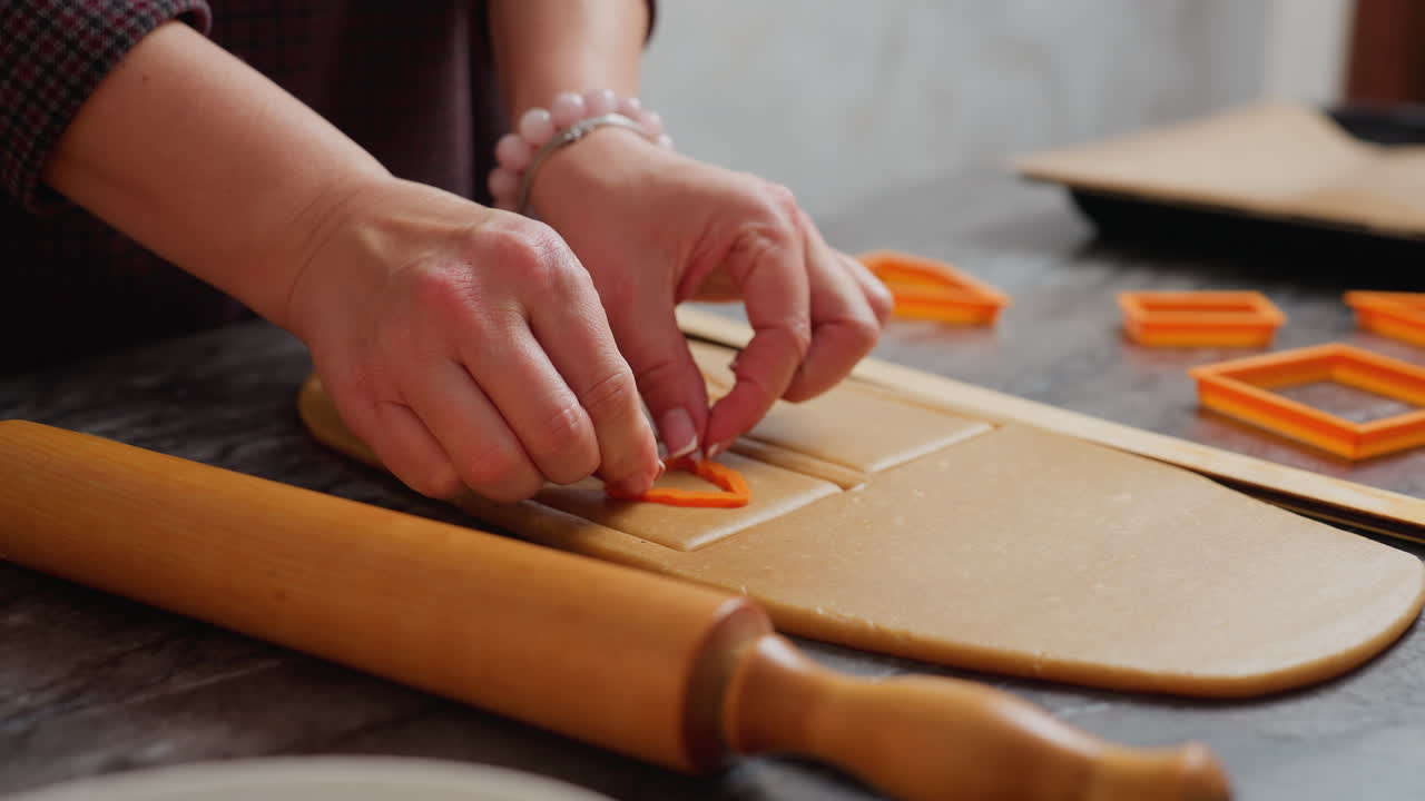 primer plano de las manos de la niña usando un cortador en forma de fresa dentro de una forma cuadrada en la masa, un rodillo de madera descansa al lado del espacio de trabajo, con otros cortadores de galletas naranjas esparcidos en el fondo