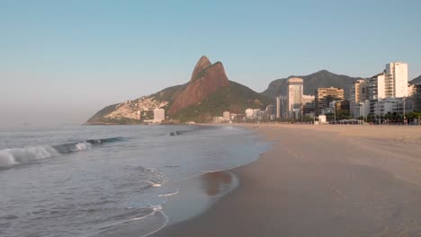 vista aérea baja hacia atrás y hacia arriba de las olas del océano que llegan a la playa vacía de la ciudad costera de río de janeiro durante la hora dorada de la madrugada