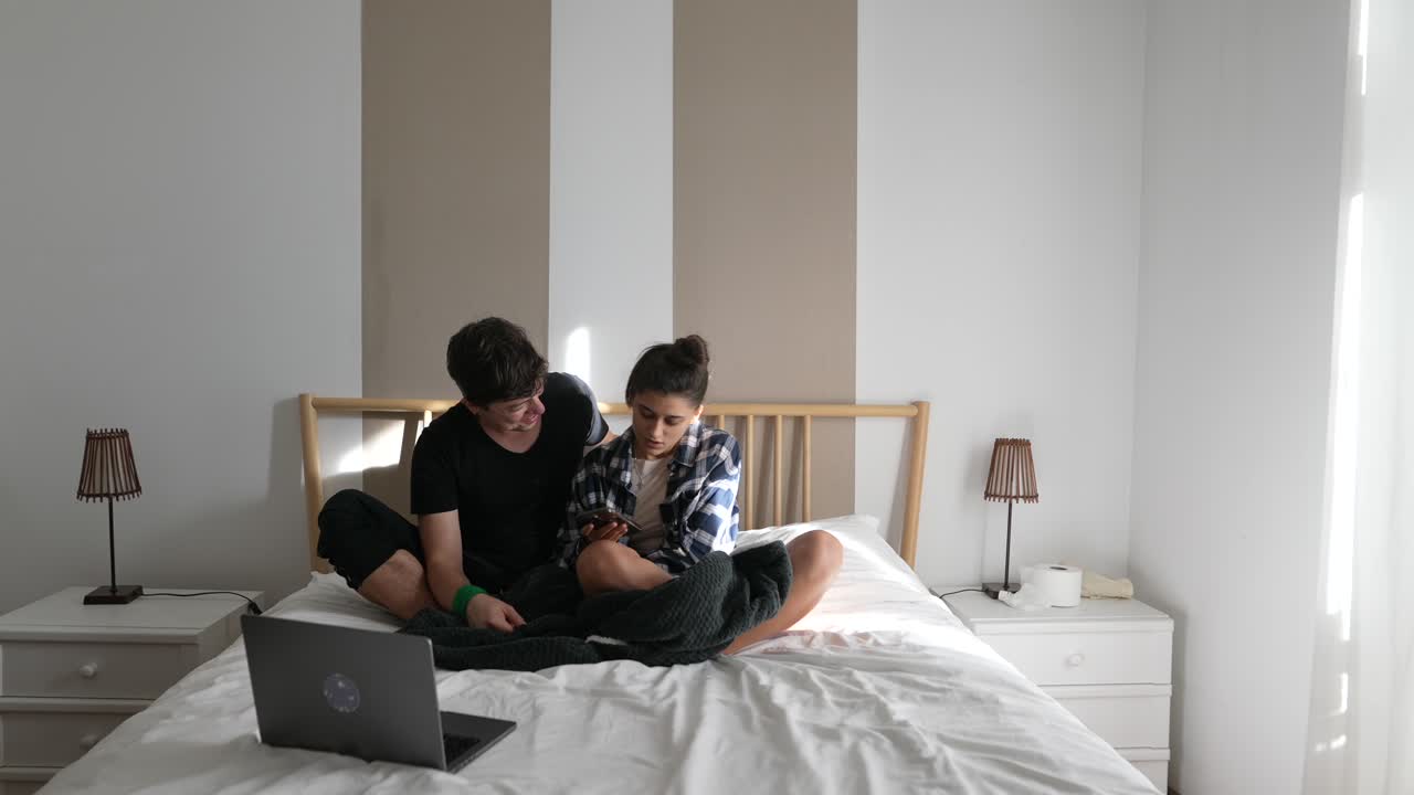 Couple using phone and laptop on a bed