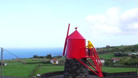 Hombre-En-El-Molino-De-Viento-Tradicional-Y-Rojo-En-La-Costa-De-La-Isla-De-Faial,-Azores,-Portugal