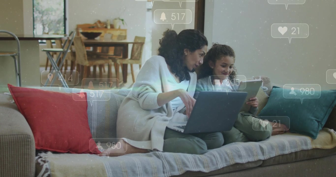 Mor med bærbar computer ser sociale ikoner dukke op, læner sig frem og peger på en tablet, mens hun vejleder sin datter.
