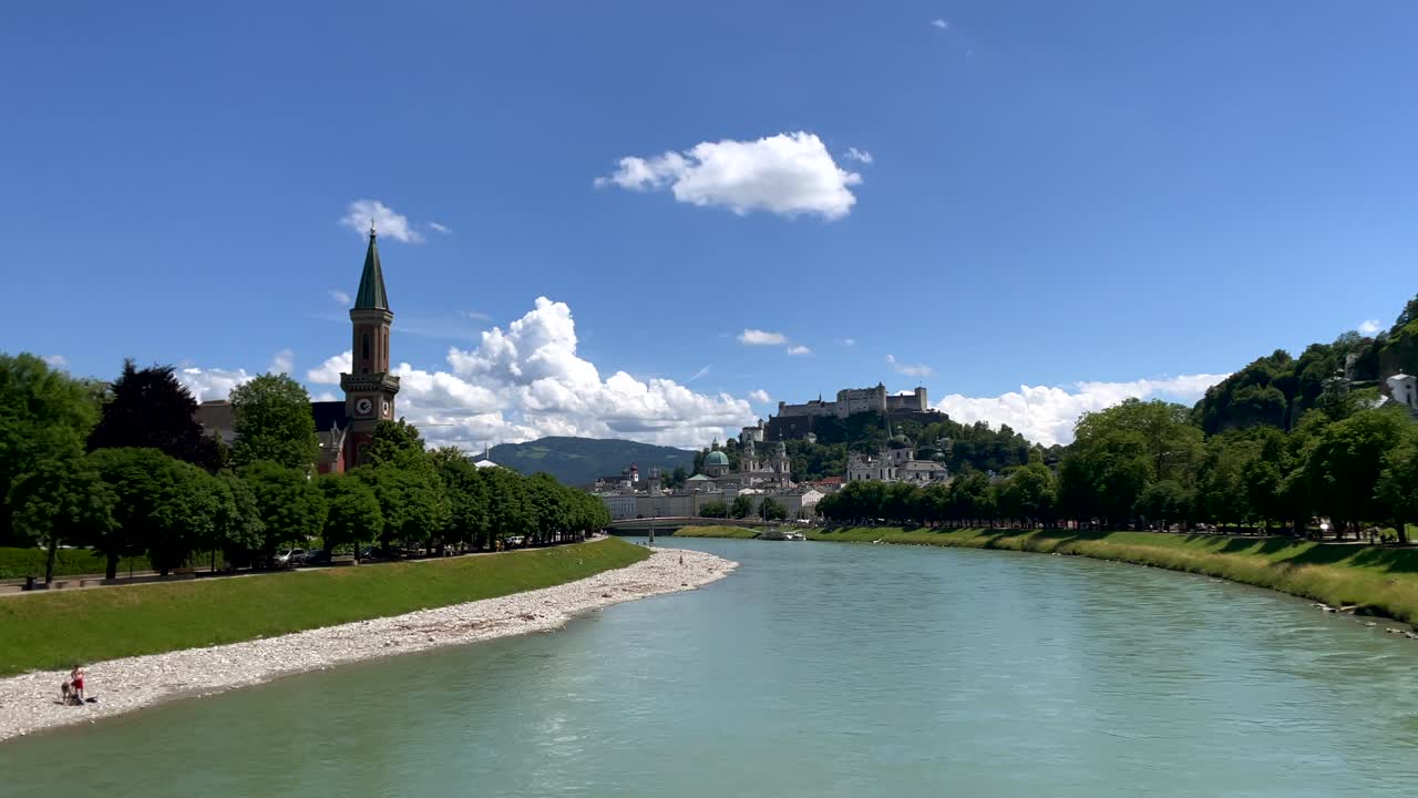 toma panorámica del río verde salzach y la ciudad austriaca de salzburgo en el fondo durante un día soleado con cielo azul - orilla rocosa del río y avenida de árboles verdes con edificios históricos en el fondo