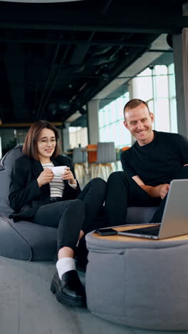 smiling woman and man look at laptop sitting in bean bag chairs. lady drinks coffee listening to her colleague. vertical video.