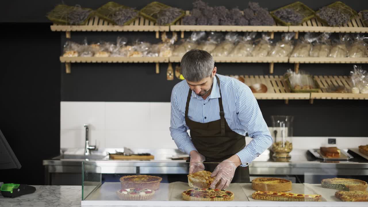 Young handsome man in the apron and gloves bringing fresh pies to the counter in the bakery shot. Indoors