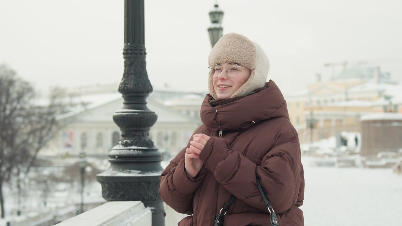 Donna in piedi a mezzo busto su un ponte urbano innevato, mani giunte in vita e un lieve sorriso che suggerisce serena contentezza, avvolta in un cappotto marrone imbottito e un cappello di maglia foderato in pelliccia con il respiro leggero visibile