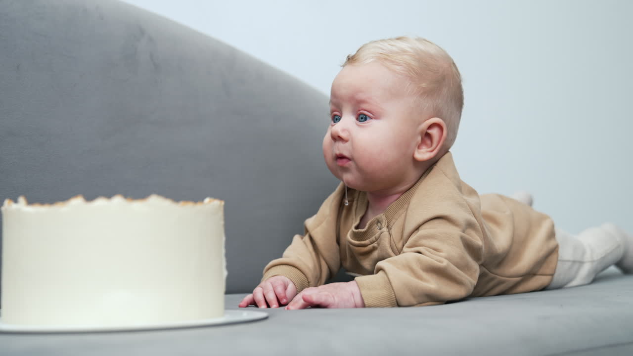 Cute blond baby with plump cheeks lies on the sofa. Kid looks at the cake in front of him and starts to cry.