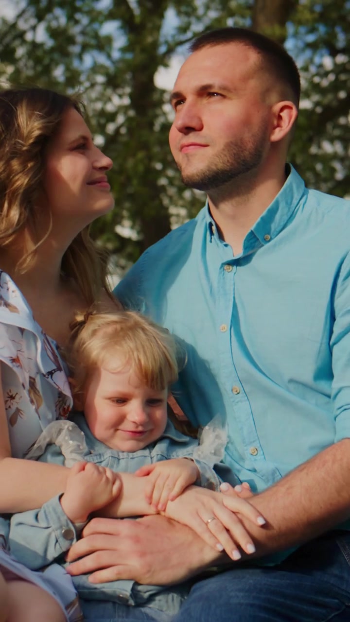 Un trio familiare sorride all'aperto sotto alberi illuminati dal sole. Mamma e papà tengono il bambino stretto, con gli occhi dolci di affetto. La scena cattura un legame familiare pacifico e gioioso immerso nella natura. Ideale per temi legati alla famiglia e alla genitorialità.