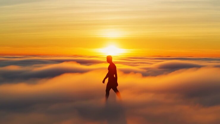 Silhouetted person walks gently above cloud sea at sunrise, centered beneath bright sun and warm golden sky, with soft haze, radiant light, long shadow, and serene uplifting atmosphere over