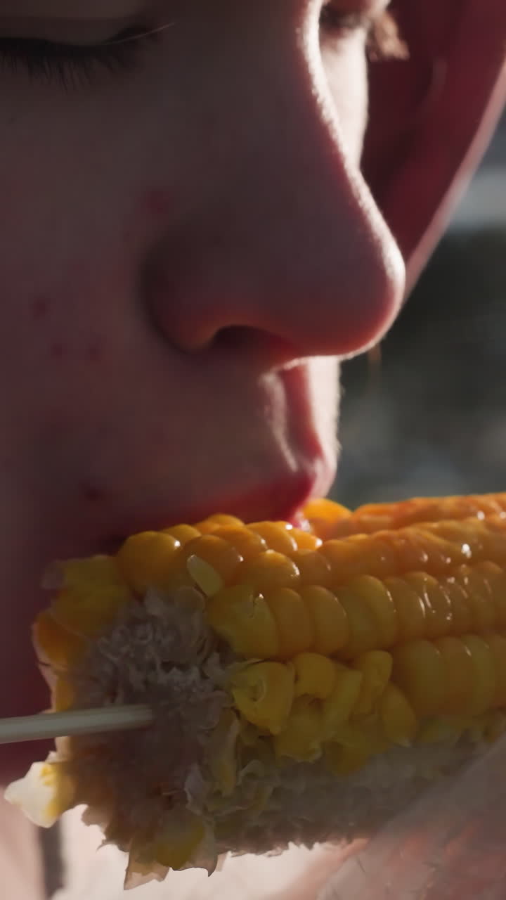 primer plano de una persona comiendo maíz sostenido por un palo envuelto en plástico blanco con luces bokeh en un fondo borroso, vapor que se eleva del maíz fresco en un entorno al aire libre