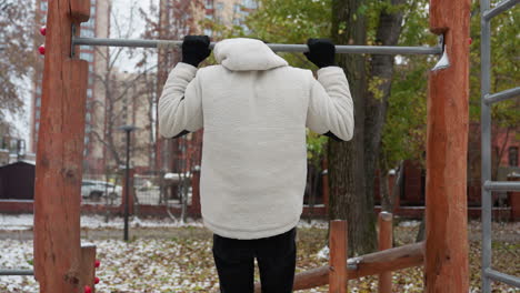 student leaps to grasp workout equipment, performing pull-ups and immediate drops among falling dry leaves, set against an urban backdrop with buildings, cars, and bare trees