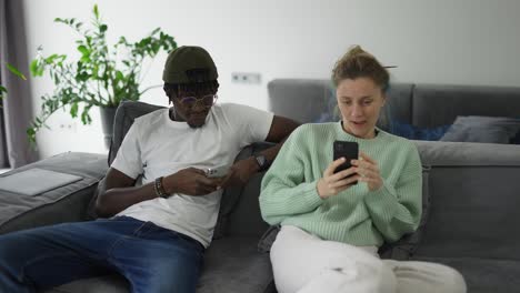 happy biracial couple relaxing on sofa with smartphones