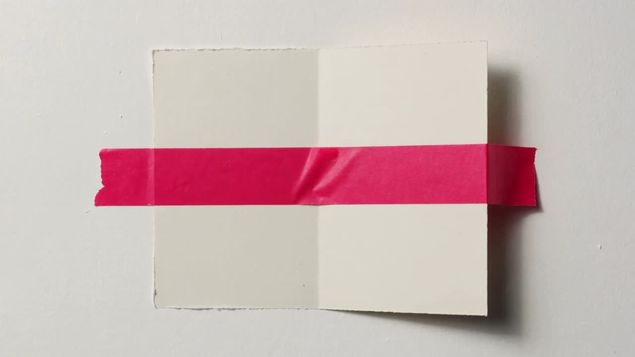Colando fita rosa num cartão dobrado sobre uma mesa branca, com os painéis a assentar rente para remover vincos.