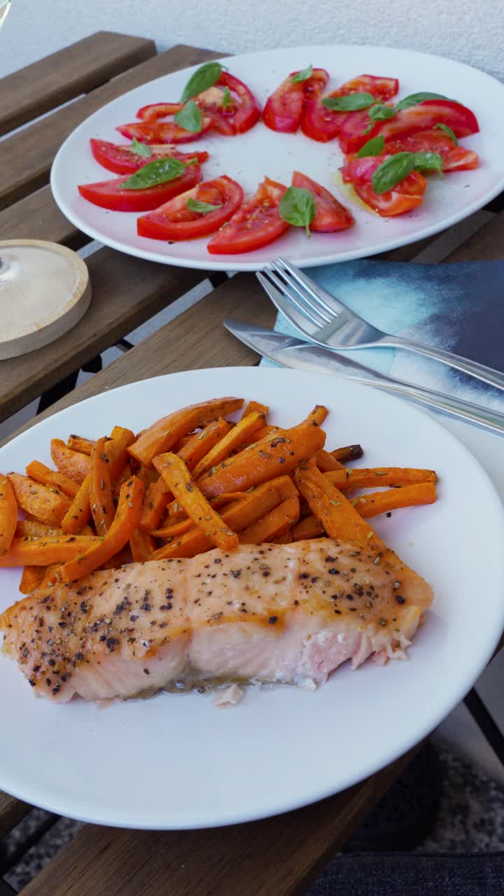 Vertical video of a plated salmon dinner with sweet potato fries and tomato basil salad served outdoors on a small balcony. Perfect for food, lifestyle, wellness, or recipe content