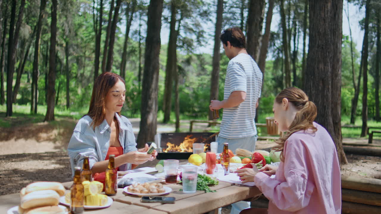 Group people preparing bbq in sunny park. Smiling two women cutting vegetables