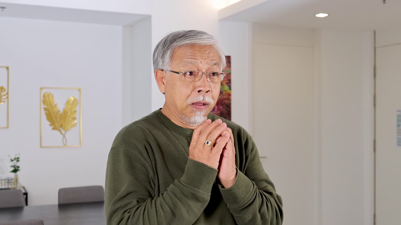 Senior man in green sweater prays sincerely in bright modern living room, soft natural lighting