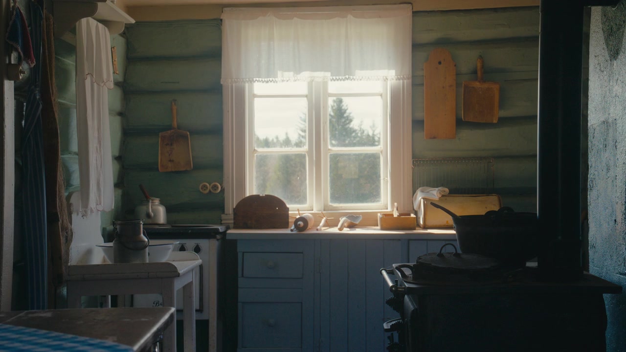 La luce soffusa del giorno filtra attraverso una finestra con tende di pizzo in una cucina vintage, dove legno consumato, utensili semplici e una stufa in ghisa creano un'atmosfera casalinga e serena.