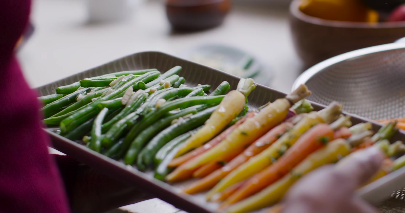 Magenta-sleeved woman lifting, tilting tray green beans and baby carrots on counter after cue
