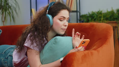 happy young woman in wireless headphones relaxing lying on sofa at home listening favorite music