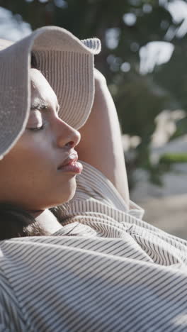 vertical video of happy biracial woman in sunhat relaxing in the sun on beach, slow motion