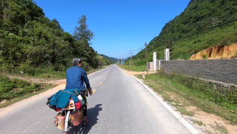 a cyclist in blue shirt riding his cycle loaded with luggage on a mountain road in cao bang province, vietnam